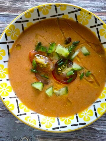 Gazpacho in a yellow and white ceramic bowl on a weathered wood table garnished with chopped tomatoes, cucumber, chives and drizzled with olive oil.