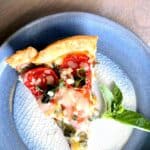A slice of tomato and cheese pie on a blue ceramic plate with a sprig of basil on the side.