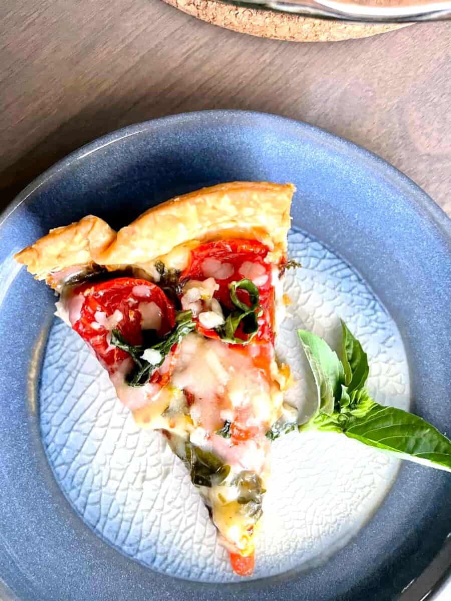 A slice of tomato and cheese pie on a blue ceramic plate with a sprig of basil on the side.