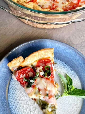 A slice of tomato and cheese pie on a blue ceramic plate with a sprig of basil on the side.