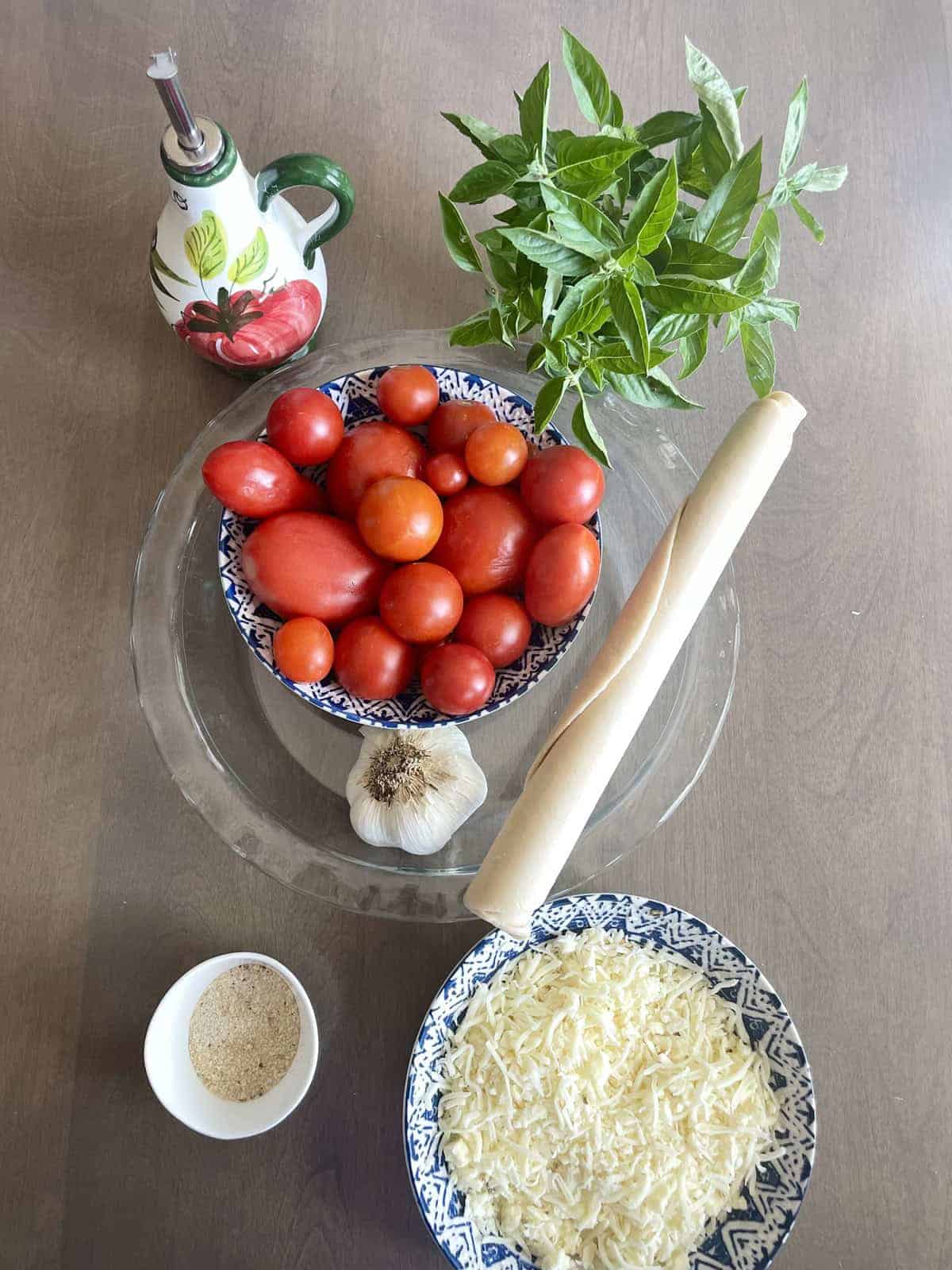 Ingredients of best southern tomato pie on a wooden table including cheese, seasoning, pie crust, garlic, tomatoes, basil, and olive oil.