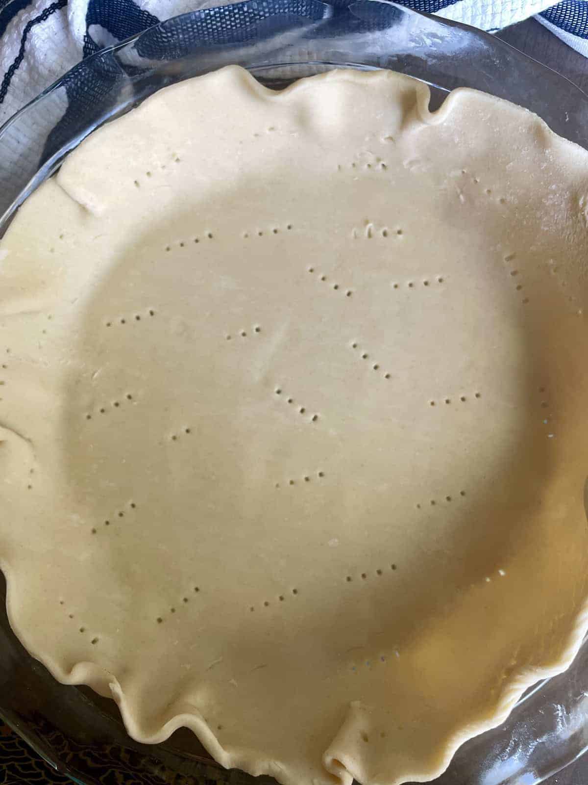 A glass pie dish filled with unbaked pie crust poked with a fork.