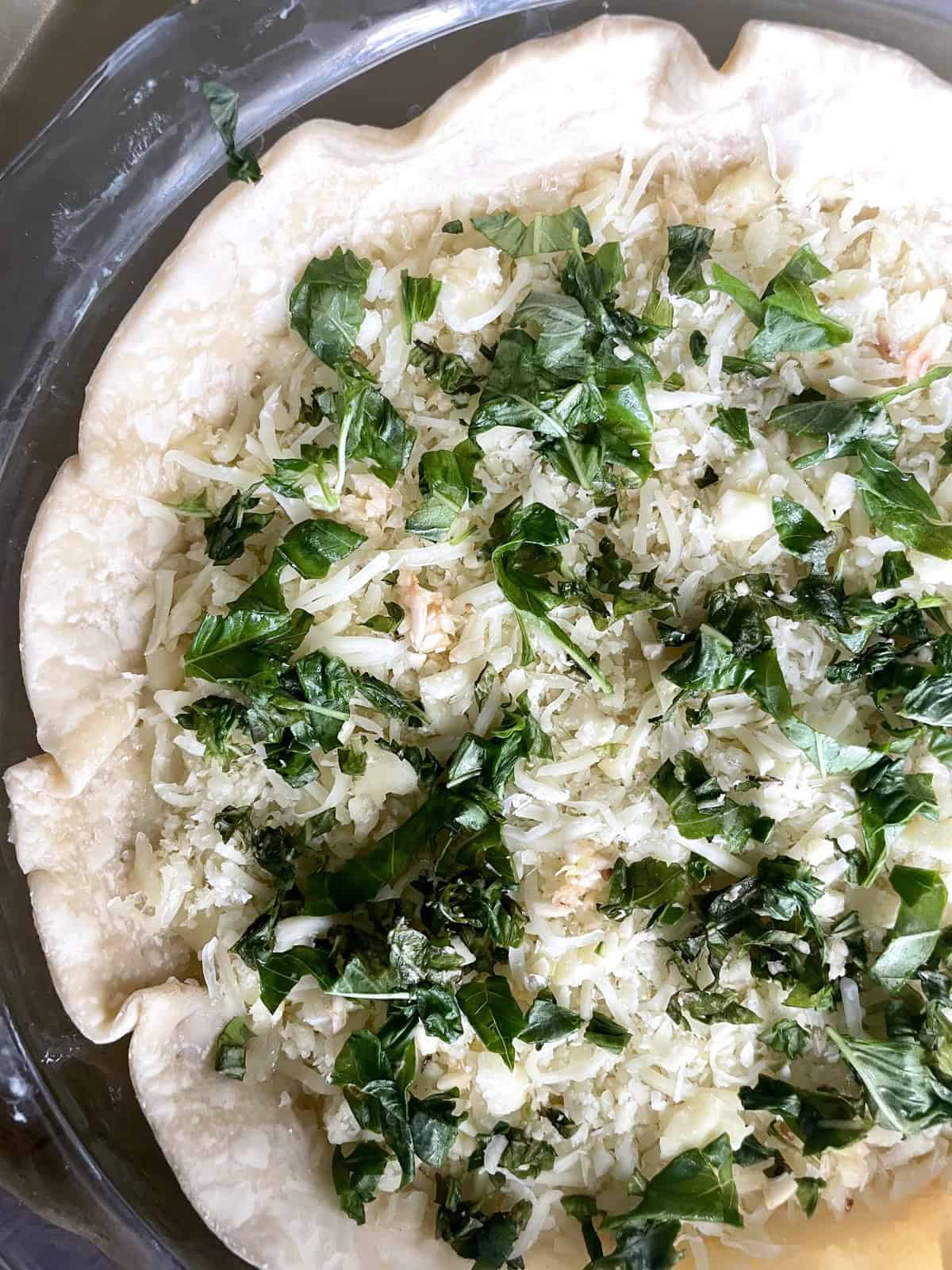 A glass pie dish filled with pie crust, grated cheese and chopped basil.