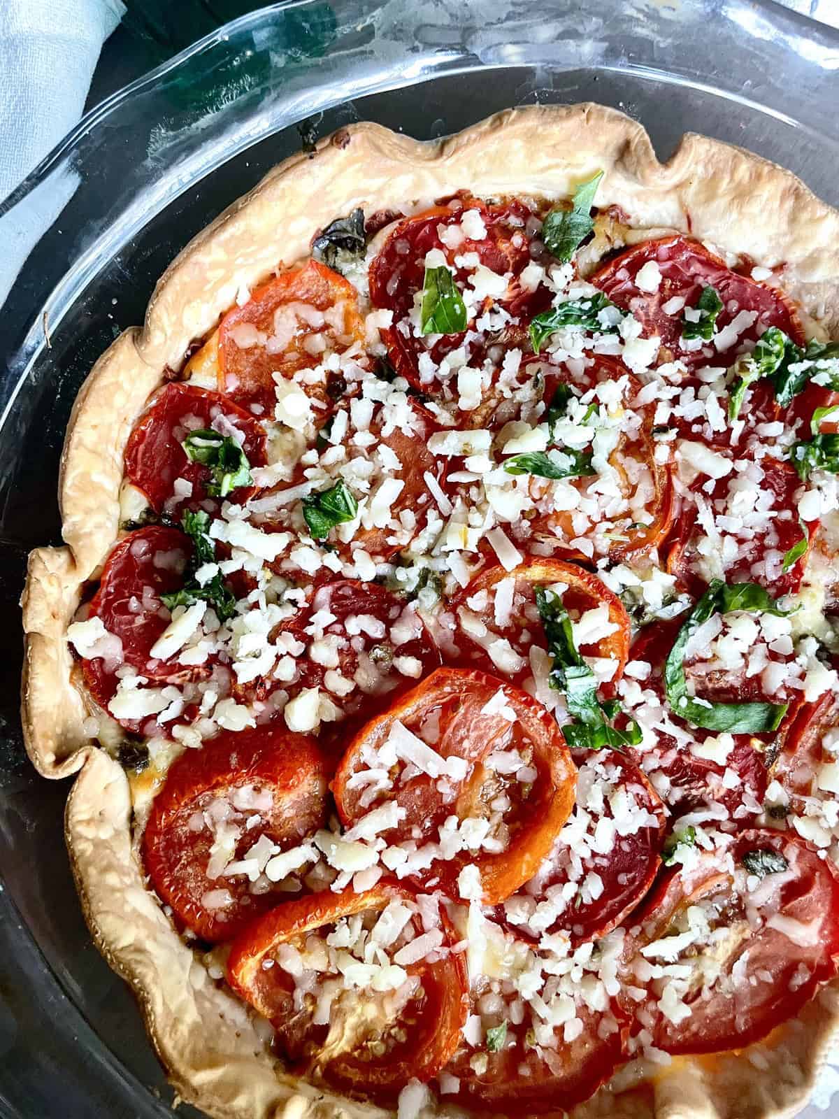 A glass pie dish filled with pie crust, sliced tomatoes, and garnished with cheese and chopped basil.