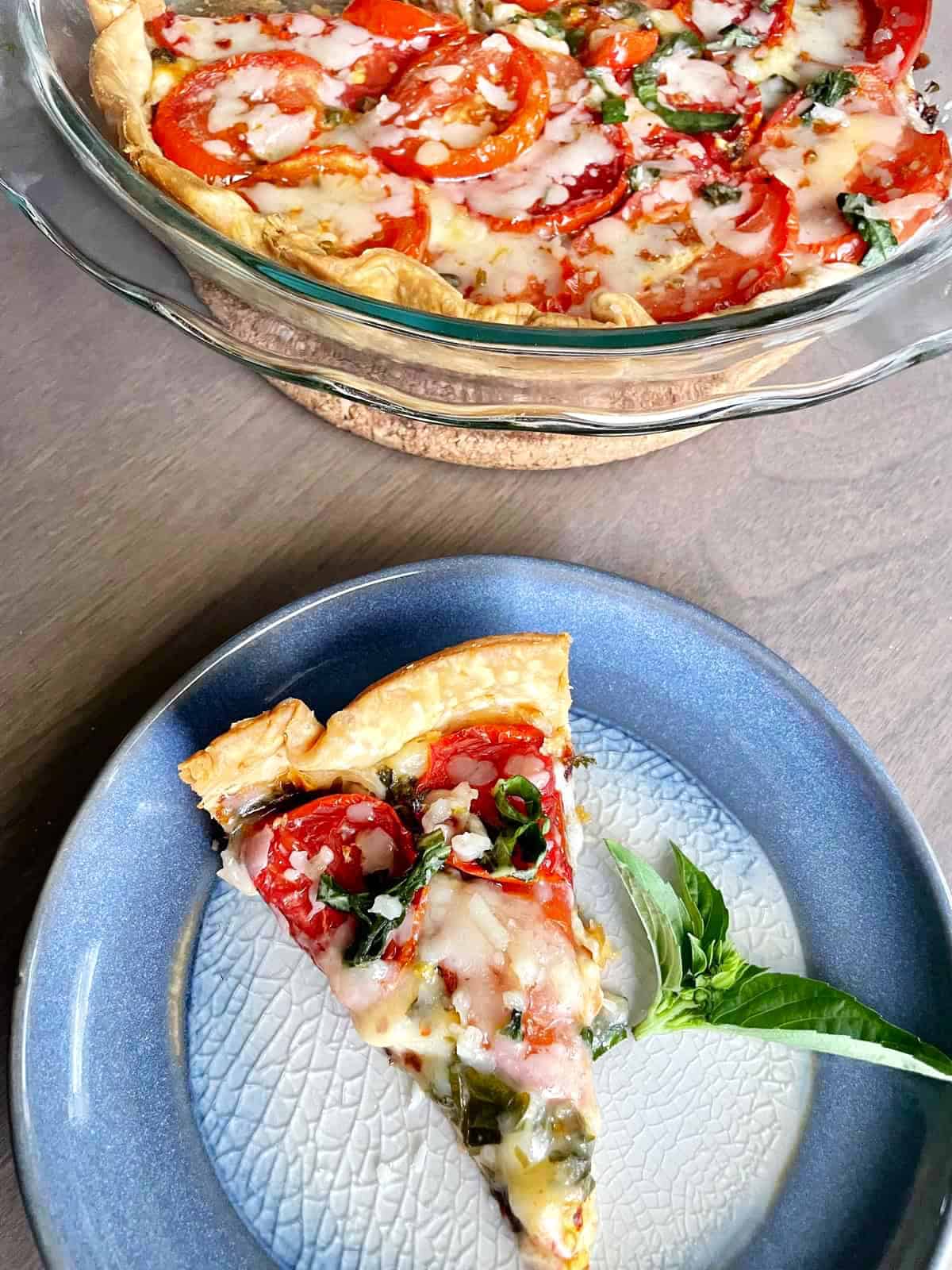 A slice of tomato and cheese pie on a blue ceramic plate with a sprig of basil on the side. A glass pie dish with the whole tomato pie is in the background.