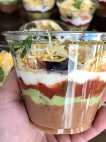 A clear cup with 7 layers of seven layer dip held in a hand.