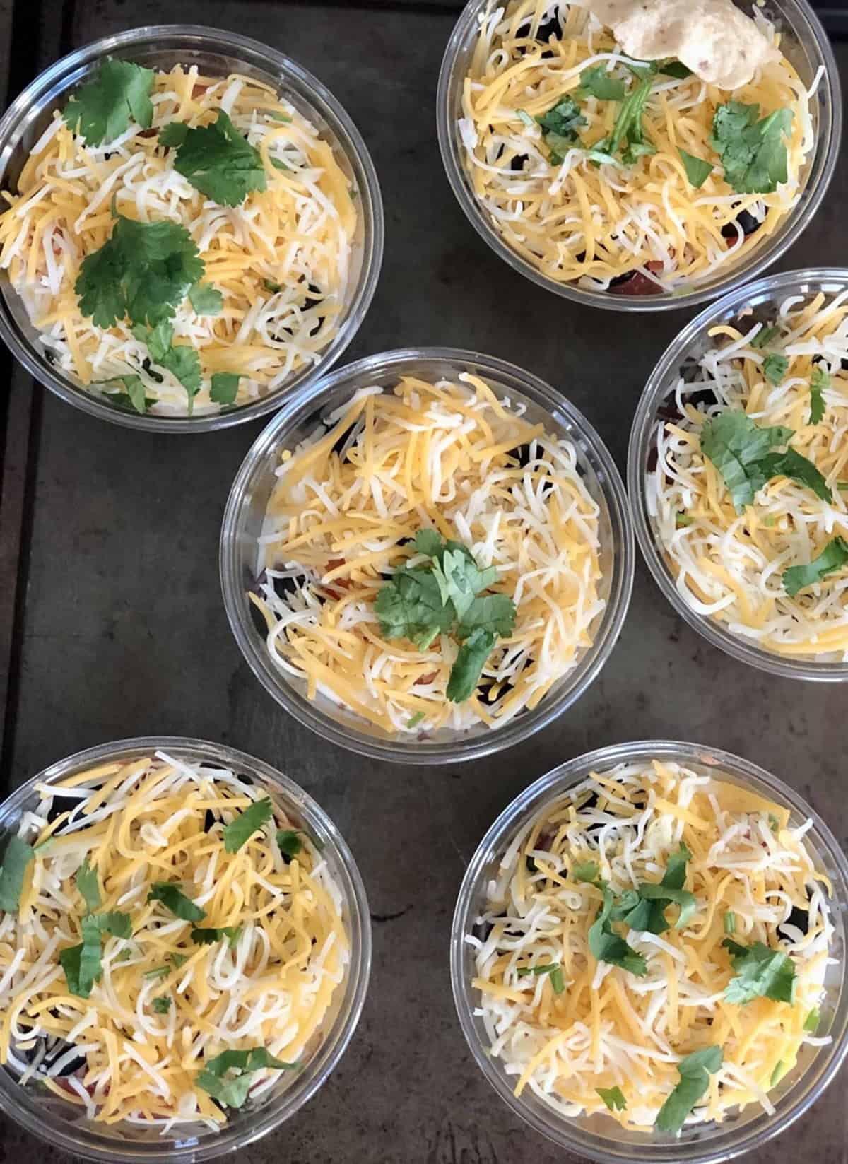 Clear cups with shredded cheese and chopped cilantro on a top on a dark metallic background.