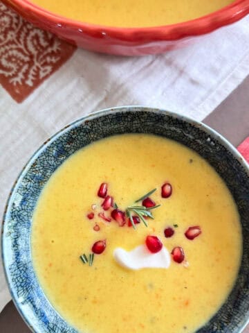 Fall apple harvest soup in a blue ceramic bowl topped with a dollop of sour cream, pomegranate, and rosemary.