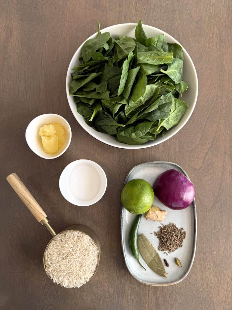 Ingredients for Indian Spinach Rice laid out on a wooden table including rice, spices, red onion, ginger, green chili, ghee, and spinach.
