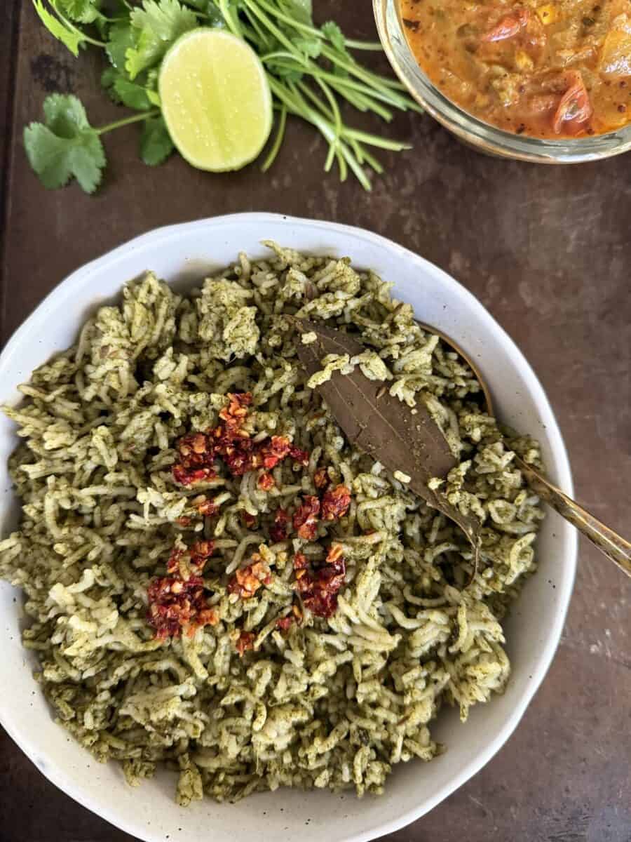 Spinach Rice with Chili crisp in a white ceramic bowl on a dark metallic background with a sliced lime, cilantro sprigs, and a bowl of curry.