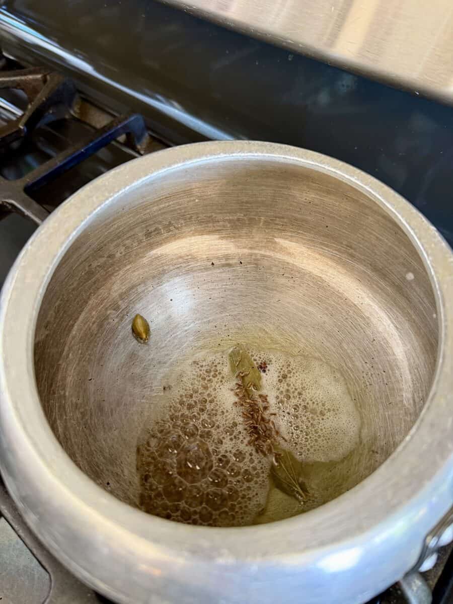 A pot with some cumin seeds, green cardamom, and bay leaf sizzling in it.