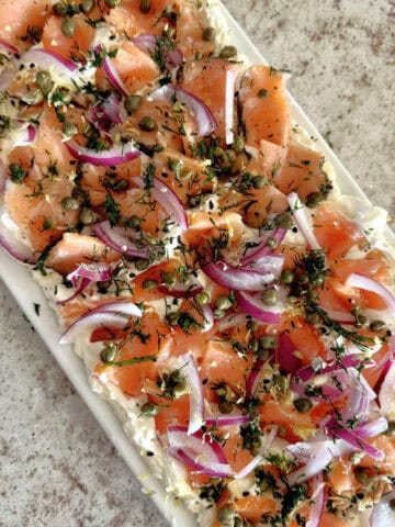 Pieces of smoked salmon, sliced red onions, and capers on a bed of cream cheese sprinkled with chopped dill on a rectangular white ceramic platter set on a white and grey countertop.