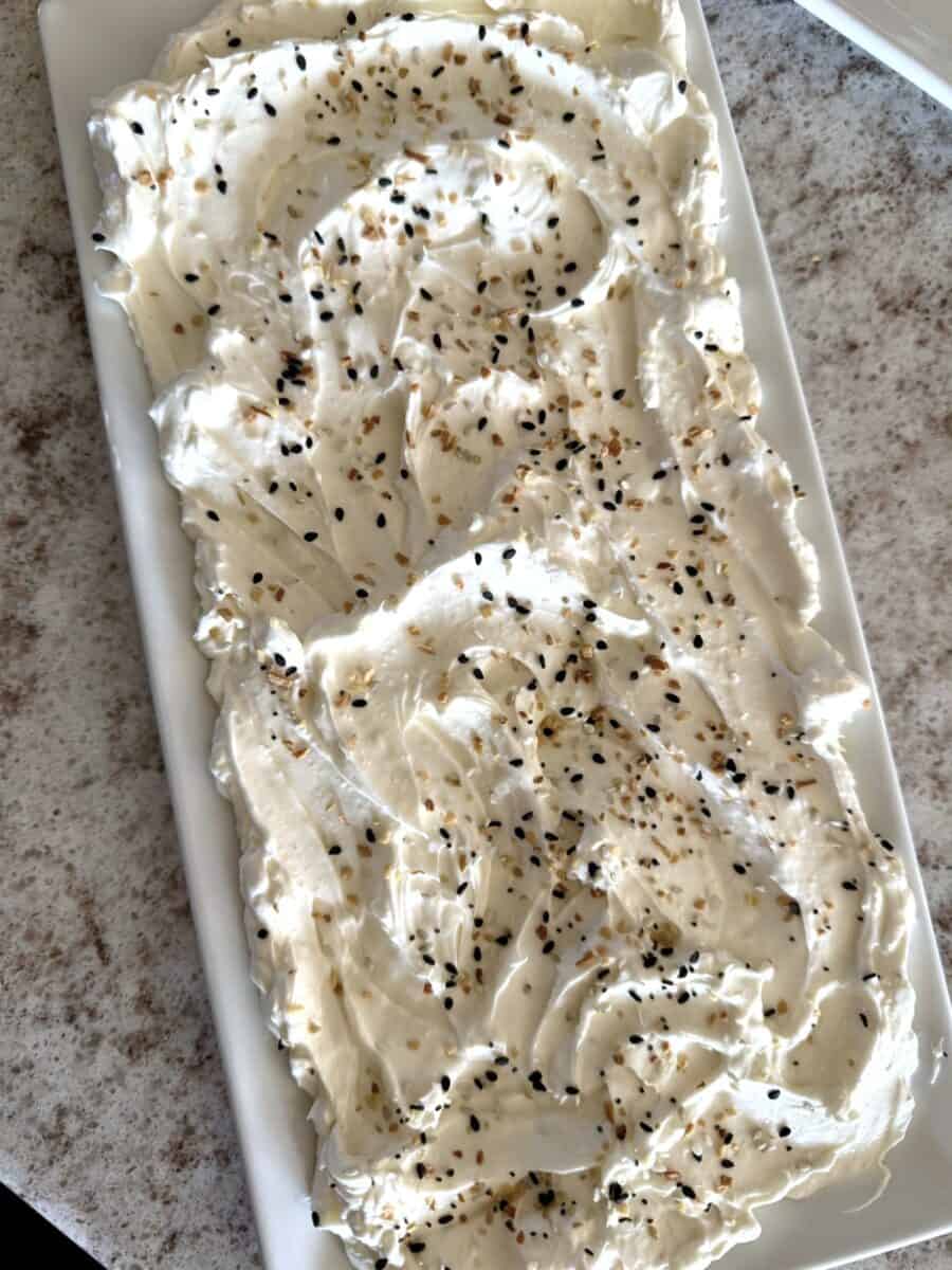 A bed of cream cheese sprinkled with everything bagel seasoning on a rectangular white ceramic platter set on a white and grey countertop.