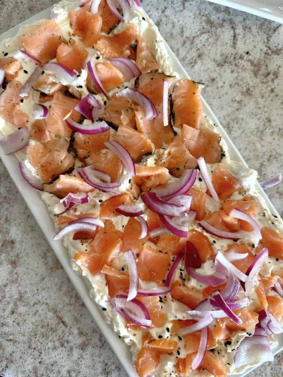 Pieces of smoked salmon and sliced red onions on a bed of cream cheese on a rectangular white ceramic platter set on a white and grey countertop.