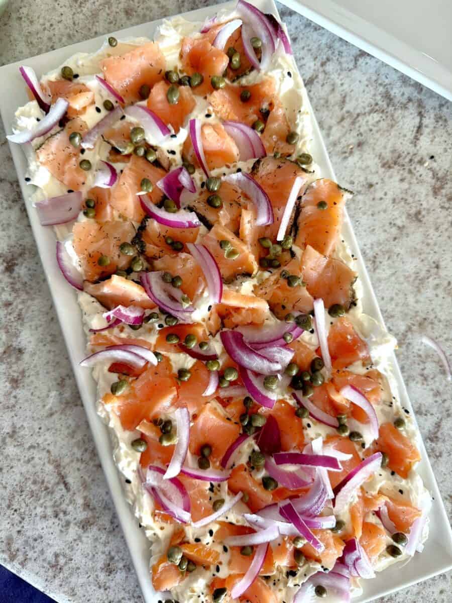 Pieces of smoked salmon, sliced red onions, and capers on a bed of cream cheese on a rectangular white ceramic platter set on a white and grey countertop.