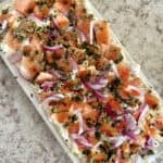 Pieces of smoked salmon, sliced red onions, and capers on a bed of cream cheese sprinkled with chopped dill on a rectangular white ceramic platter set on a white and grey countertop.
