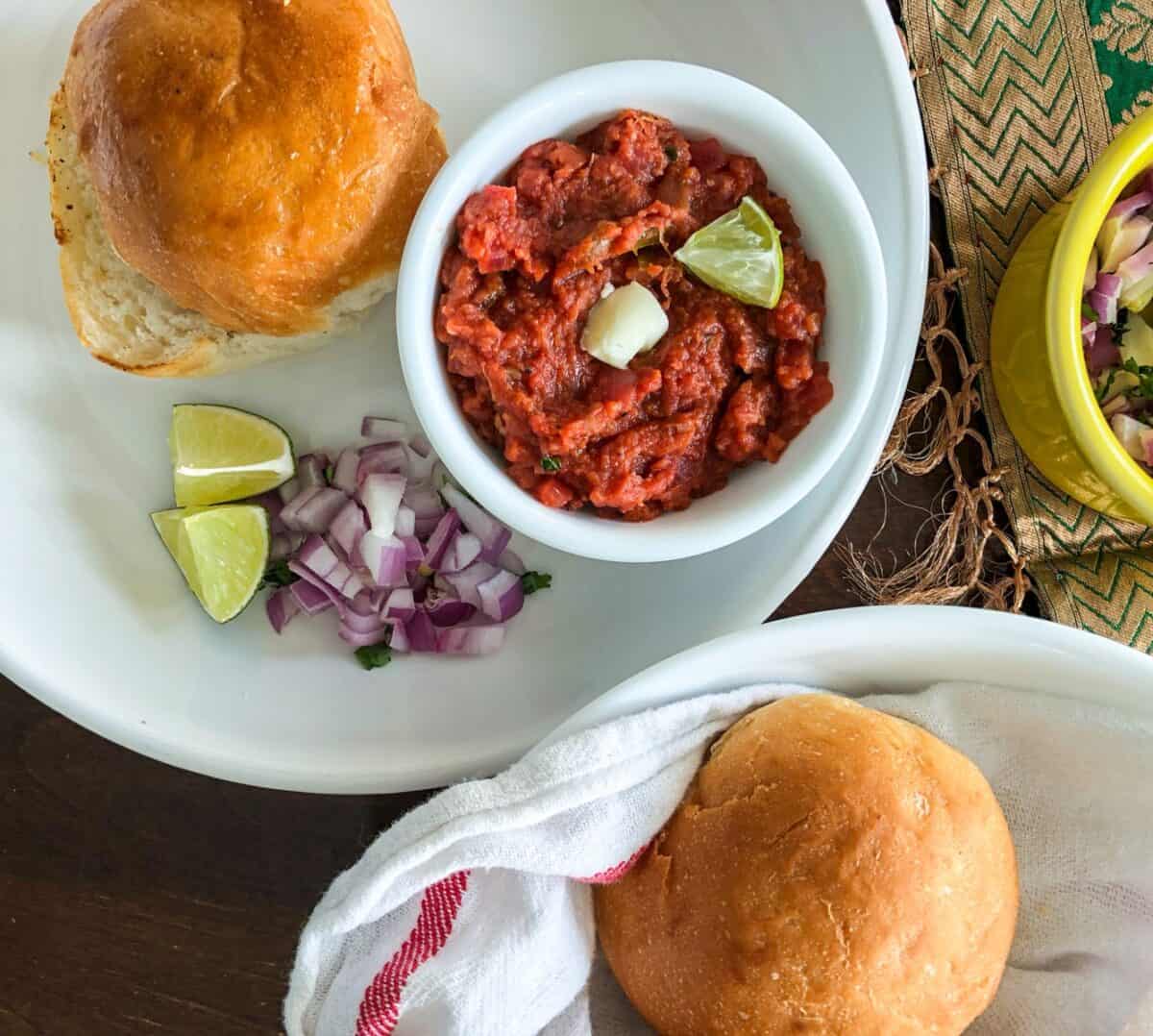 Pav, Indian dinner rolls served with a vegetable mash in a bowl, garnished with butter and a piece of lime on a plate. Chopped red onions and lime pieces are served on the side on a white ceramic plate.