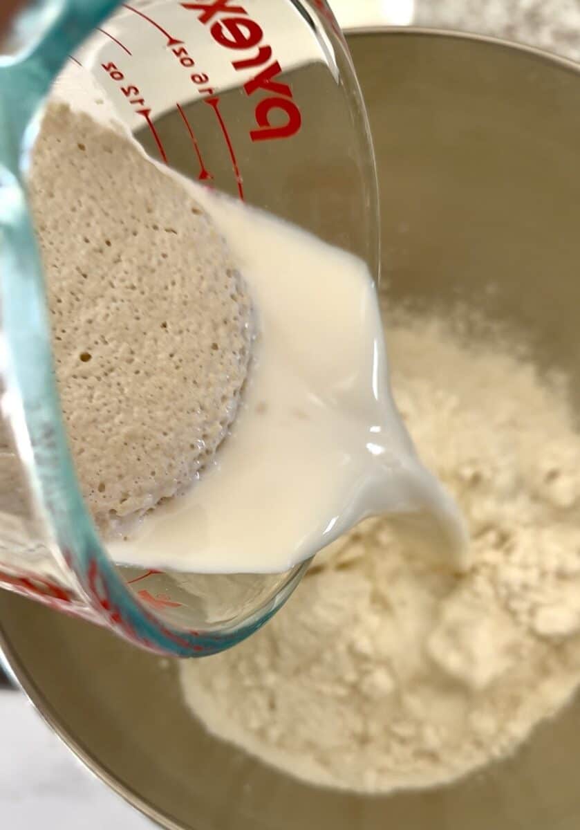 Yeast and milk mixture being poured into a stainless-steel mixing bowl with flour.