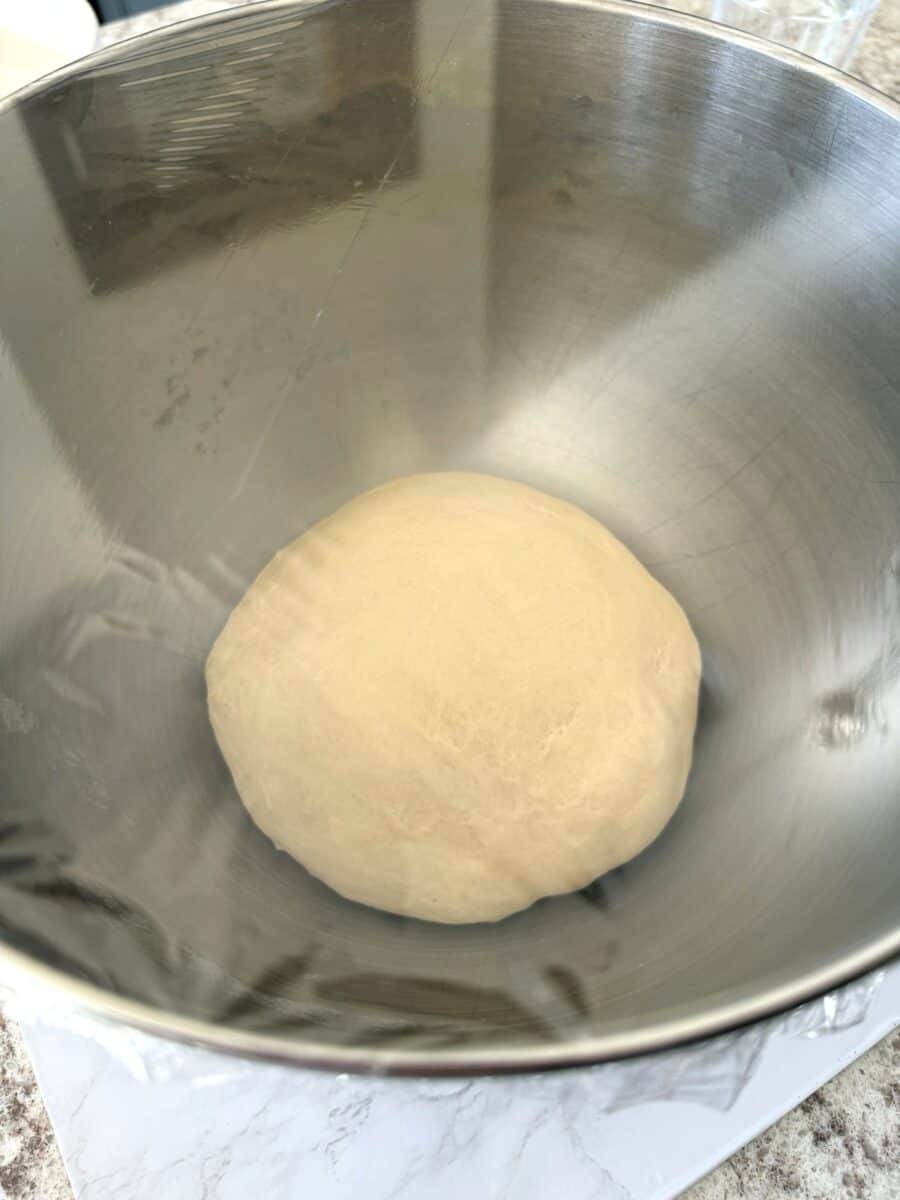 A dough ball in a stainless steel mixing bowl covered with plastic wrap.