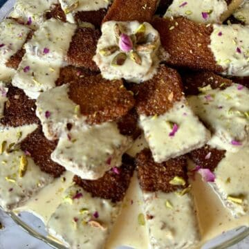 Pieces of fried bread, half dipped in golden milk and garnished with chopped pistachios and rose petals arranged on a glass cake stand.