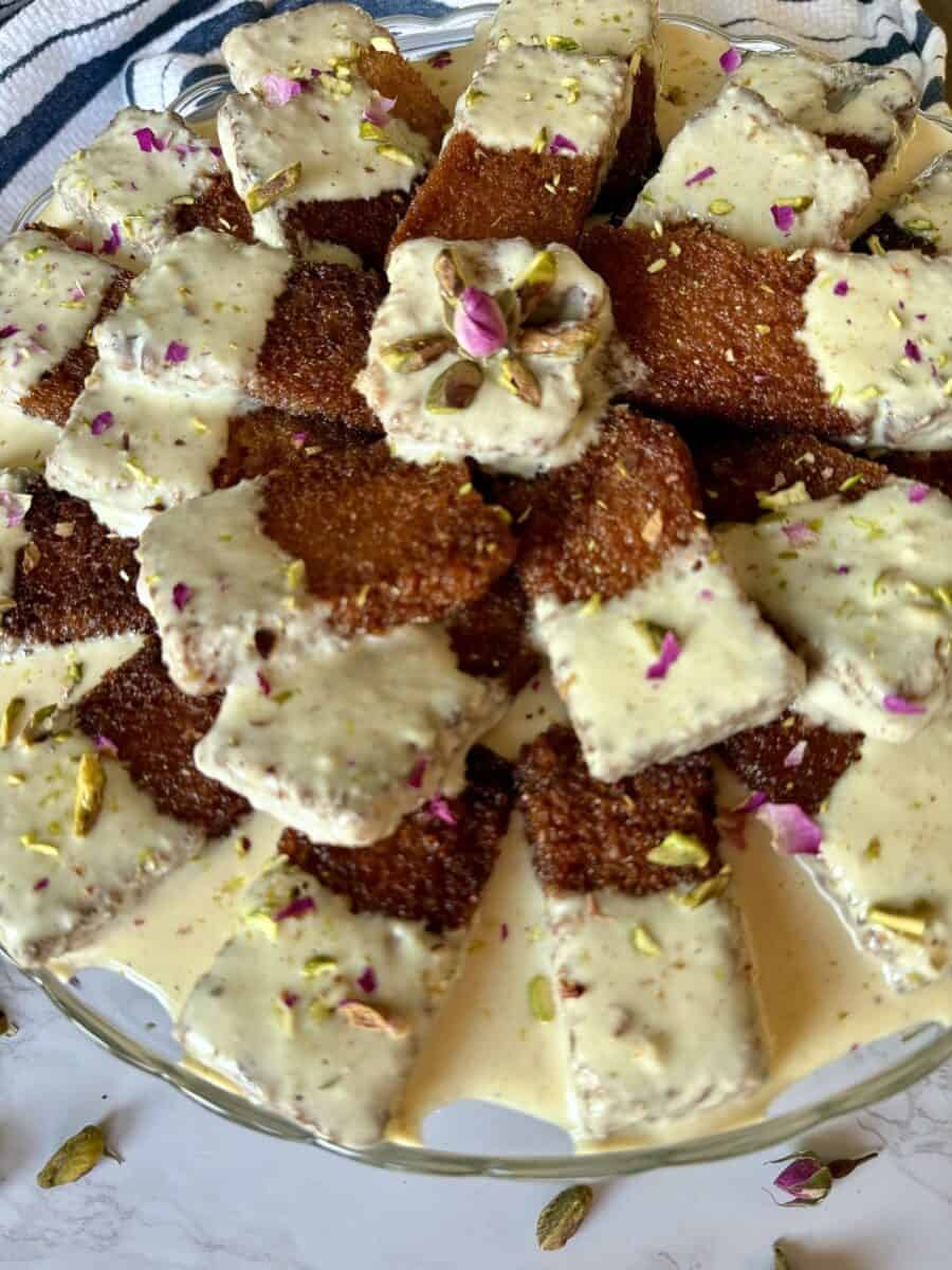 Pieces of fried bread, half dipped in golden milk and garnished with chopped pistachios and rose petals arranged on a glass cake stand.