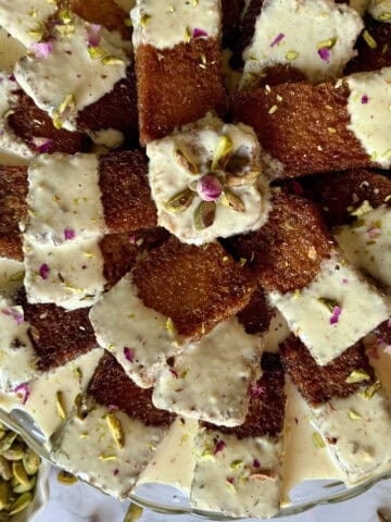 Pieces of fried bread, half dipped in golden milk and garnished with chopped pistachios and rose petals arranged on a glass cake stand.