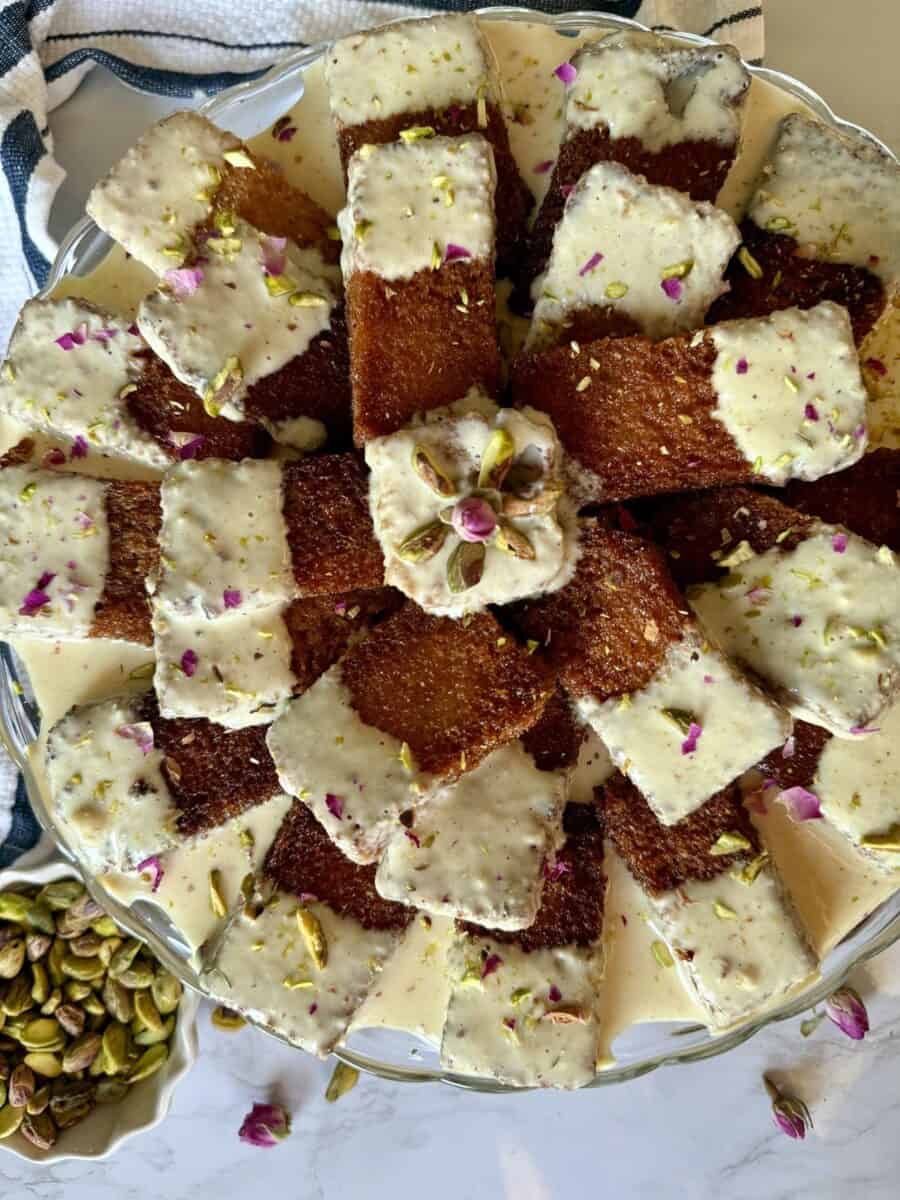 Pieces of fried bread, half dipped in golden milk and garnished with chopped pistachios and rose petals arranged on a glass cake stand.