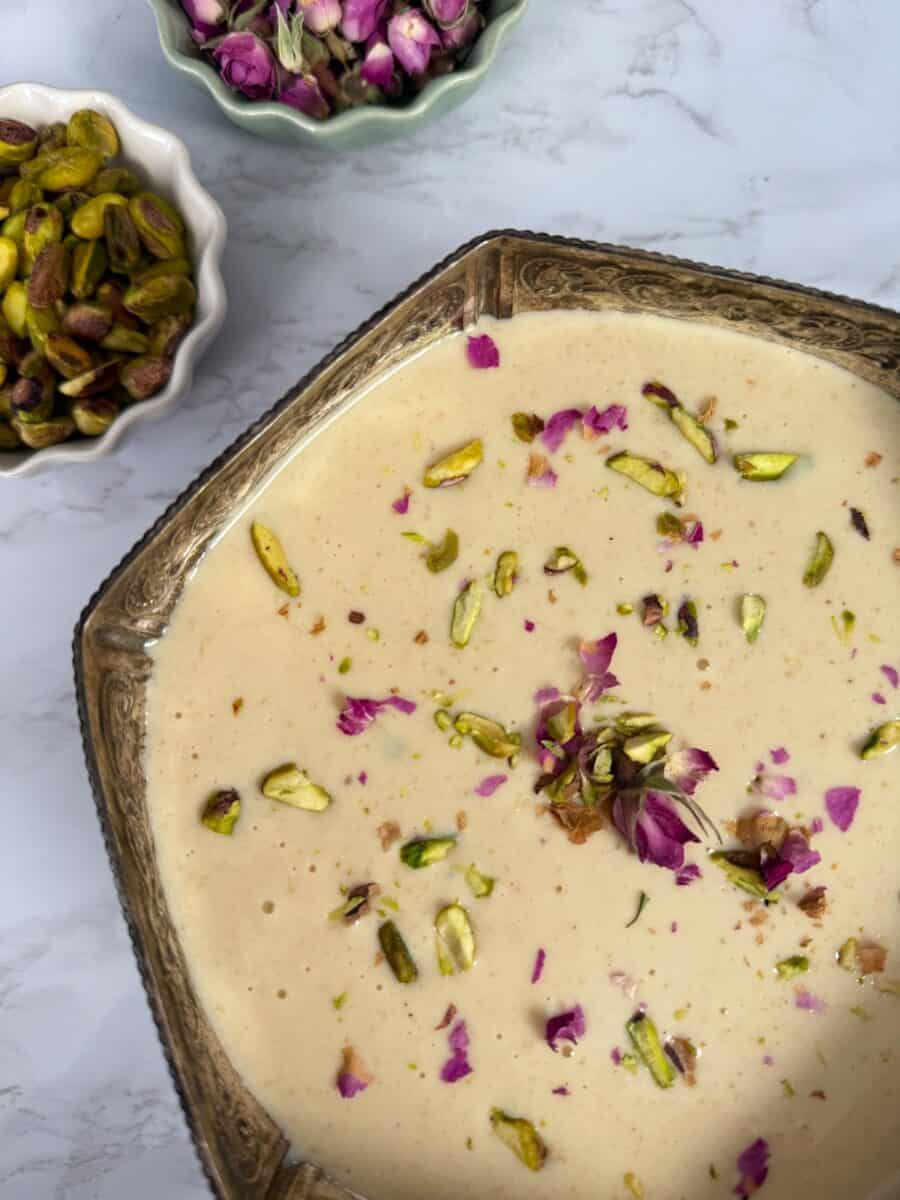 Rabdi - a preparation of milk garnished with chopped pistachios and rose petals in a silver serving bowl on a white marble background