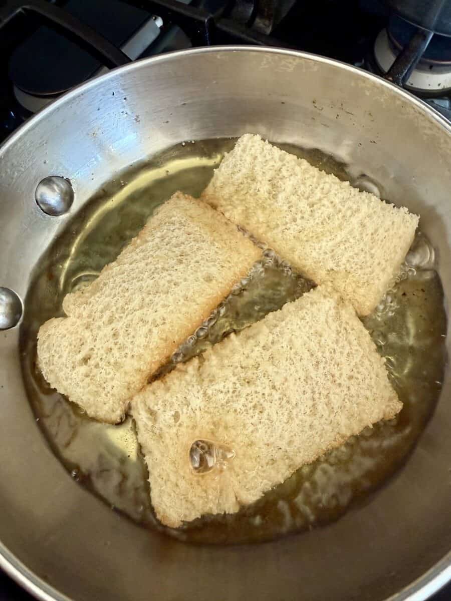 Pieces of white bread frying in oil in a stainless steel frying pan.