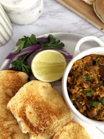 Buttered and toasted dinner rolls on a ceramic plate set on a white marble background. There is a small bowl of ground chicken Keema garnished with cilantro, a slice of lemon, and sliced red onion.