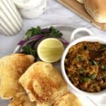 Buttered and toasted dinner rolls on a ceramic plate set on a white marble background. There is a small bowl of ground chicken Keema garnished with cilantro, a slice of lemon, and sliced red onion.