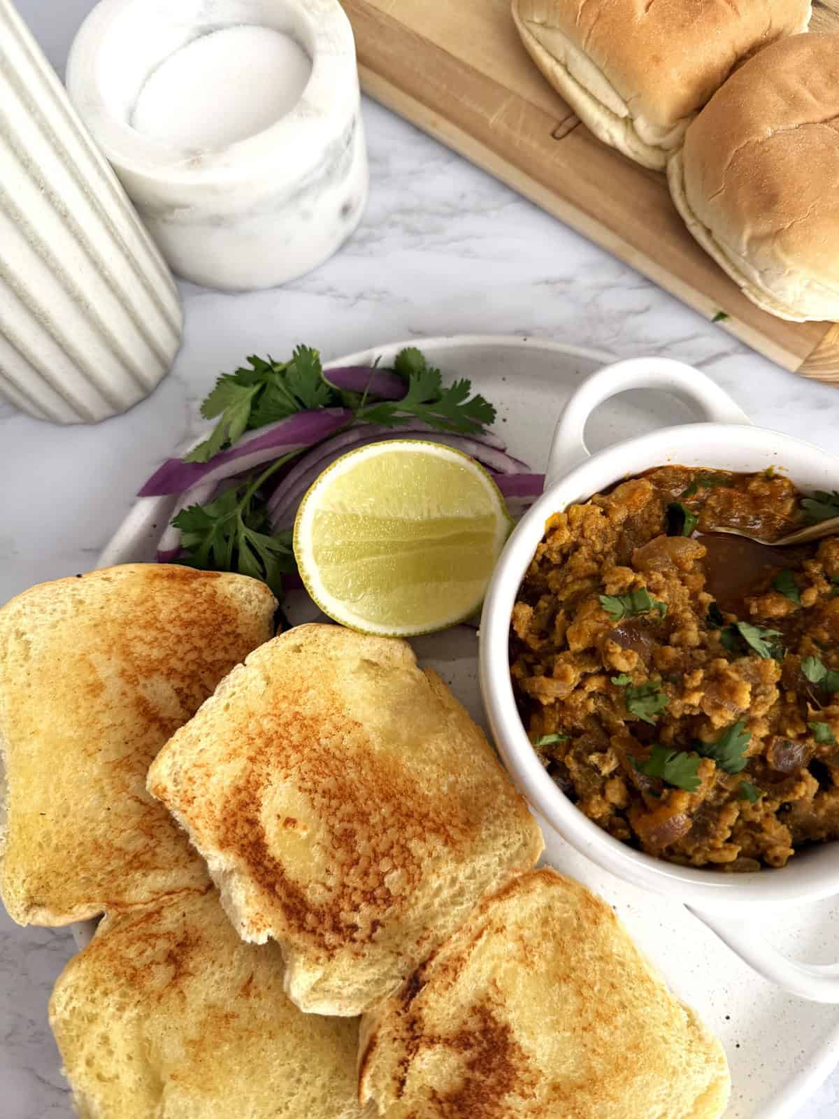 Buttered and toasted dinner rolls on a ceramic plate set on a white marble background. There is a small bowl of ground chicken Keema garnished with cilantro, a slice of lemon, and sliced red onion.