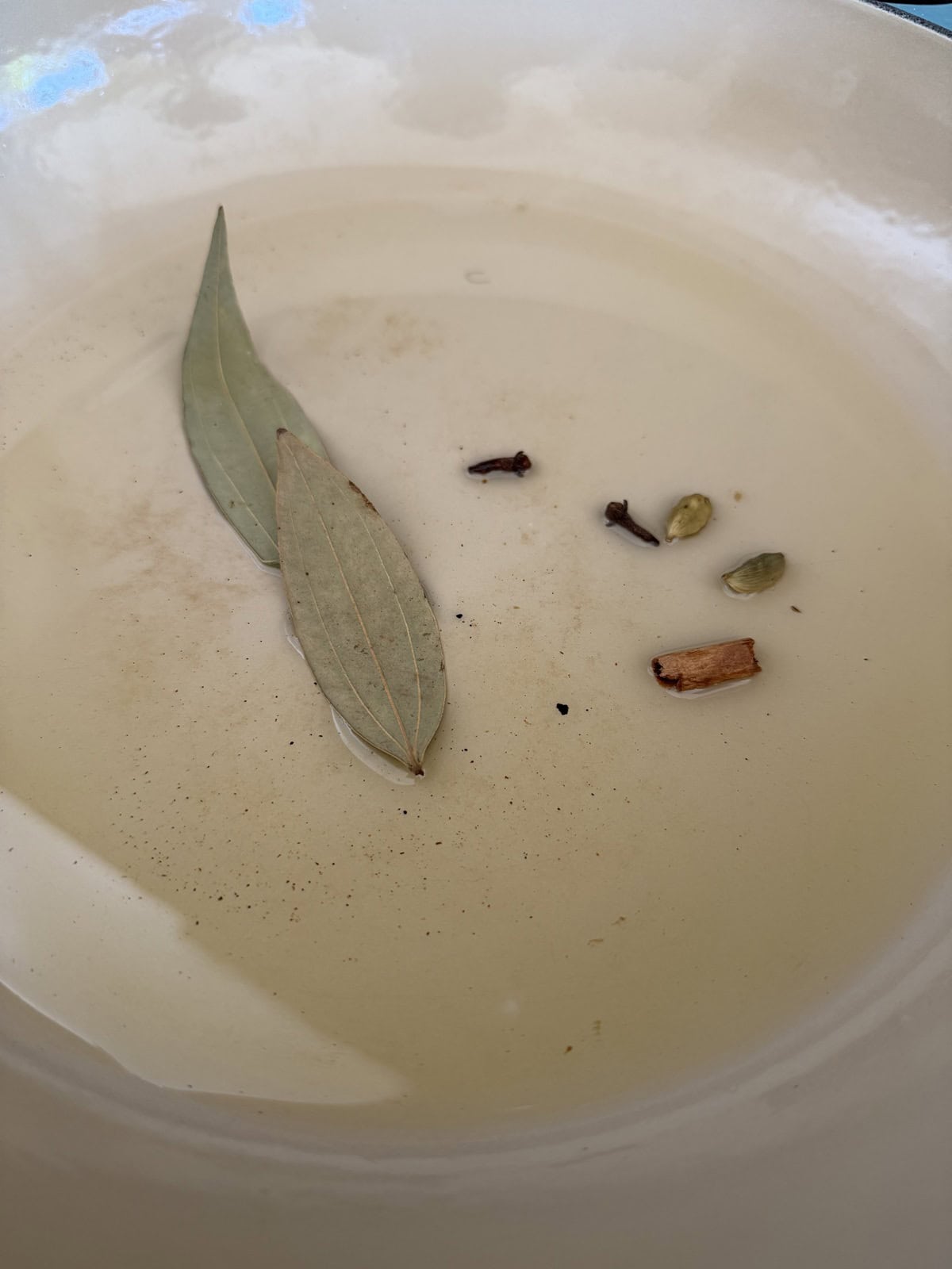 An enameled cast iron pan with oil, green cardamom, bay leaves, cloves and piece of cinnamon.