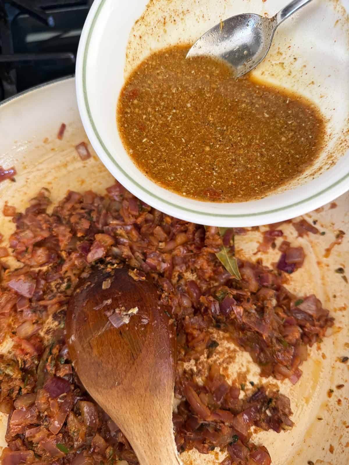 A masala of onion and tomato with spices cooking in a pan and a spice slurry being poured into it.