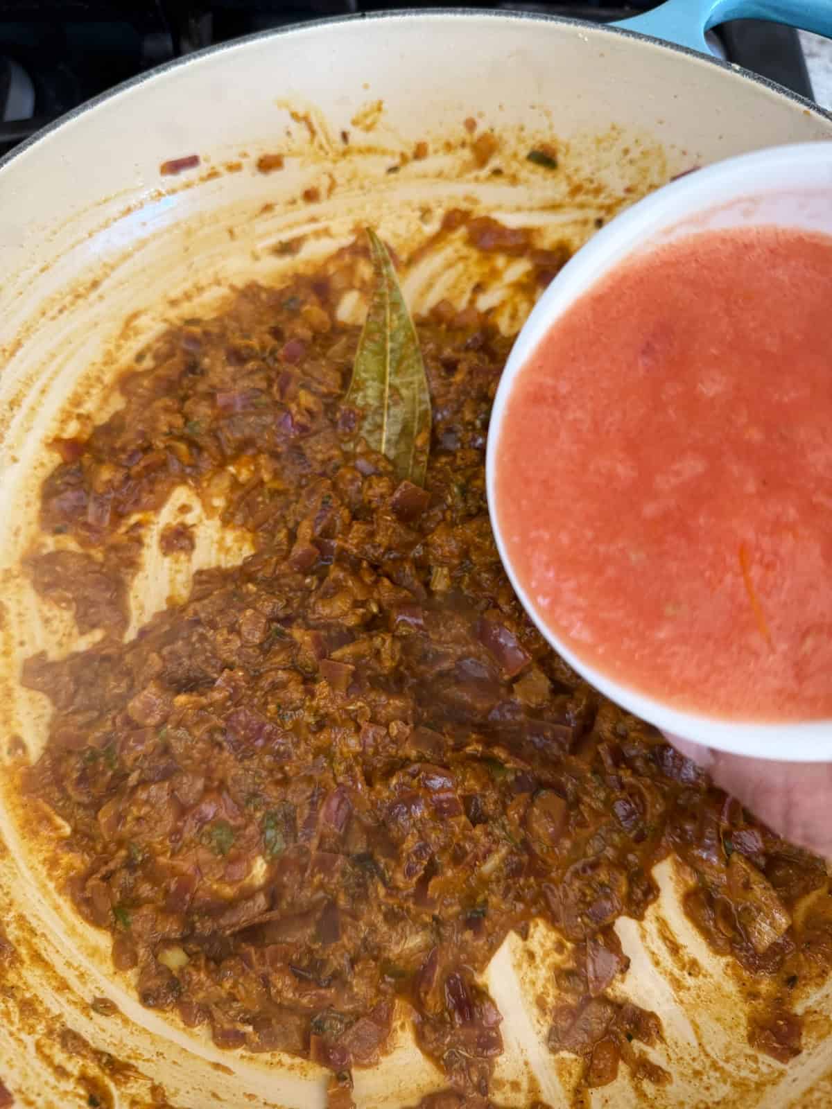 A masala of onion and tomato with spices cooking in a pan, with a bowl of tomato puree being poured into it.