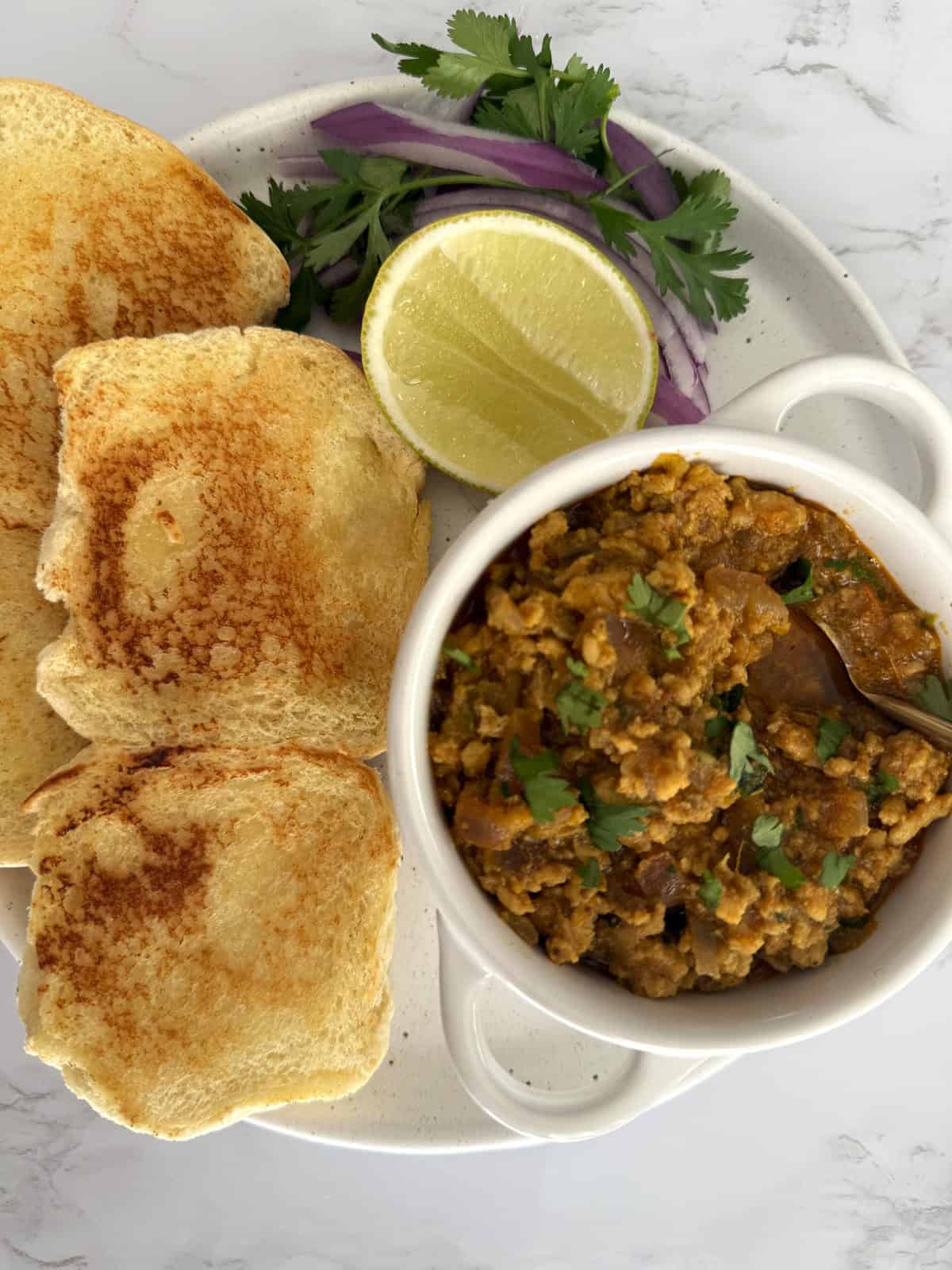 Buttered and toasted dinner rolls on a ceramic plate set on a white marble background. There is a small bowl of ground chicken Keema garnished with cilantro, a slice of lemon, and sliced red onion.