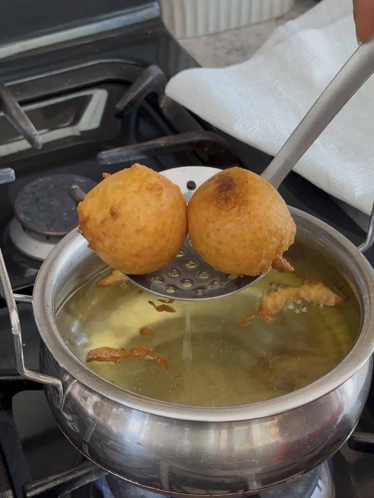 Fried food balls (mumbai batata vada) being lifted from hot oil with a skimmer in a kitchen setting.