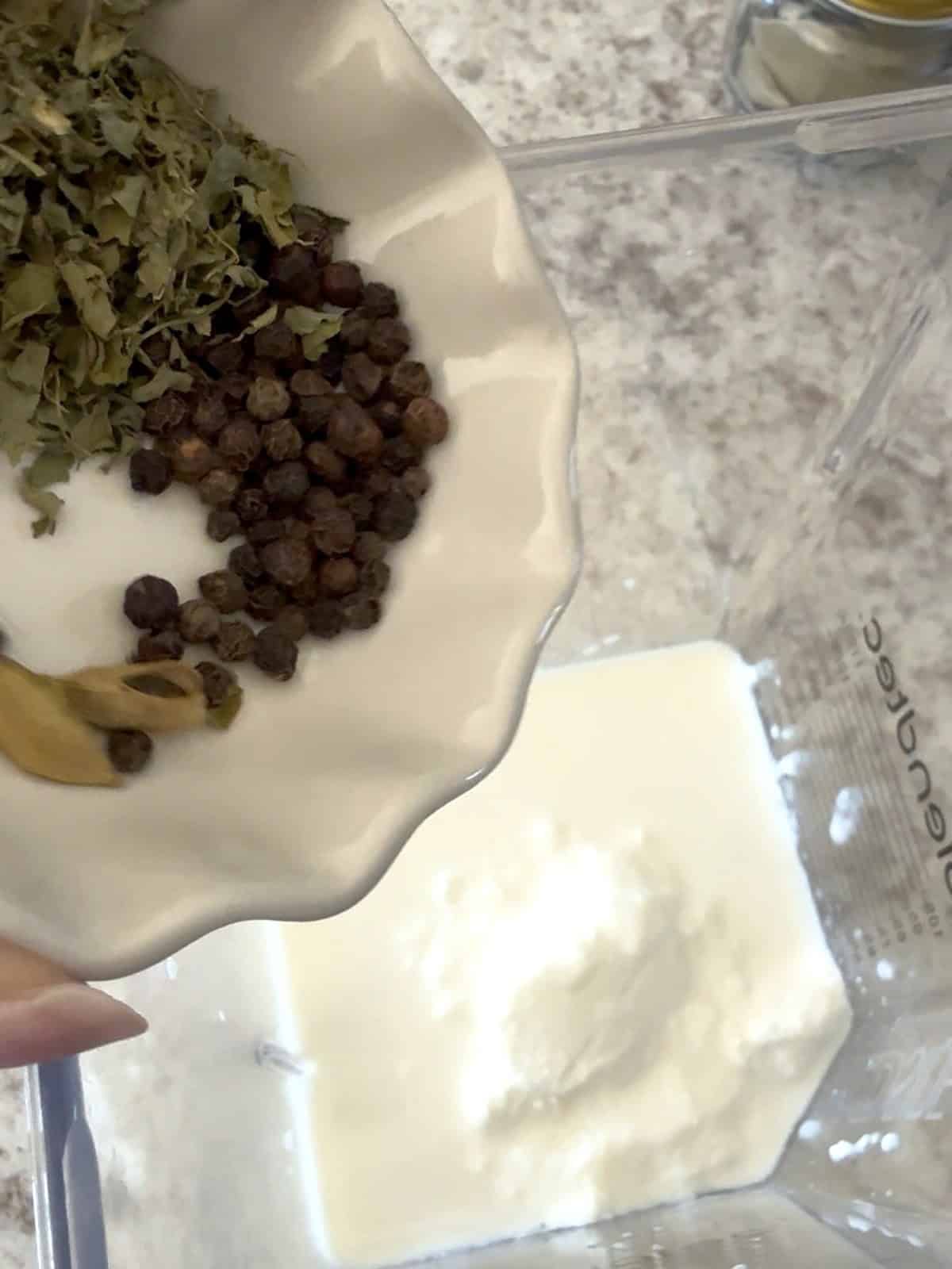 Dried fenugreek leaves, black peppercorns, and green cardamom are being added to a blender jar with heavy cream and yogurt to make a blended marinade for Afghani Chicken.