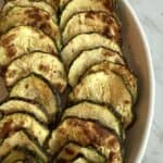 Air fried crispy zucchini slices sprinkled with chaat masala in a white dish on a white marble background.