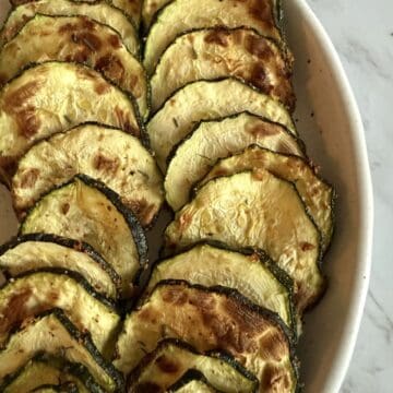 Air fried crispy zucchini slices sprinkled with chaat masala in a white dish on a white marble background.