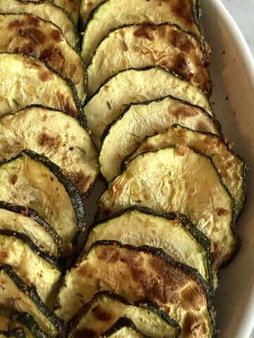 Air fried crispy zucchini slices sprinkled with chaat masala in a white dish on a white marble background.