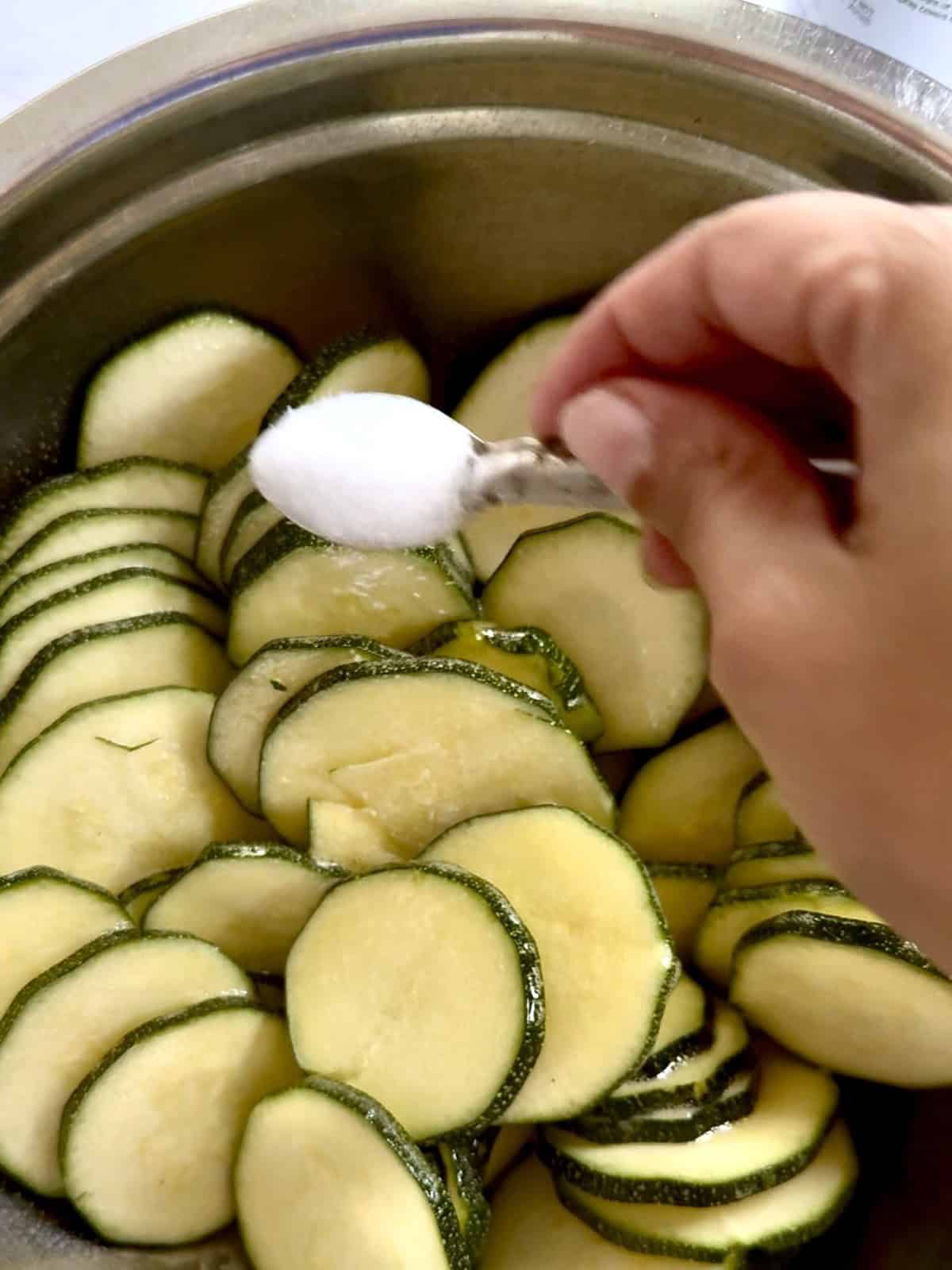 Step-by-step process for sweating water out of zucchini slices with salt. Zucchini slices in a stainless steel bowl.