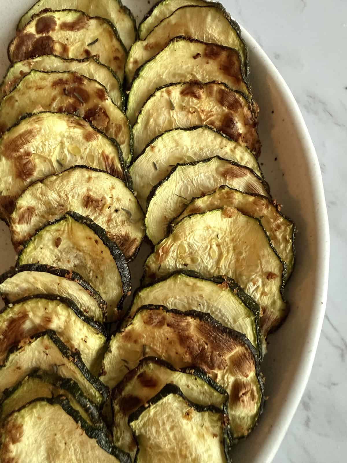 Air fried crispy zucchini slices sprinkled with chaat masala in a white dish on a white marble background.
