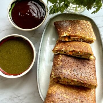 High-protein moong dal cheela with cottage cheese and tofu on a ceramic platter with mint and tamarind chutneys in two bowls on a white marble background.
