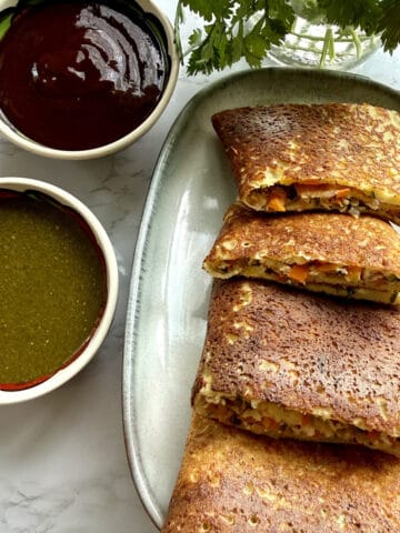 High-protein moong dal cheela with cottage cheese and tofu on a ceramic platter with mint and tamarind chutneys in two bowls on a white marble background.