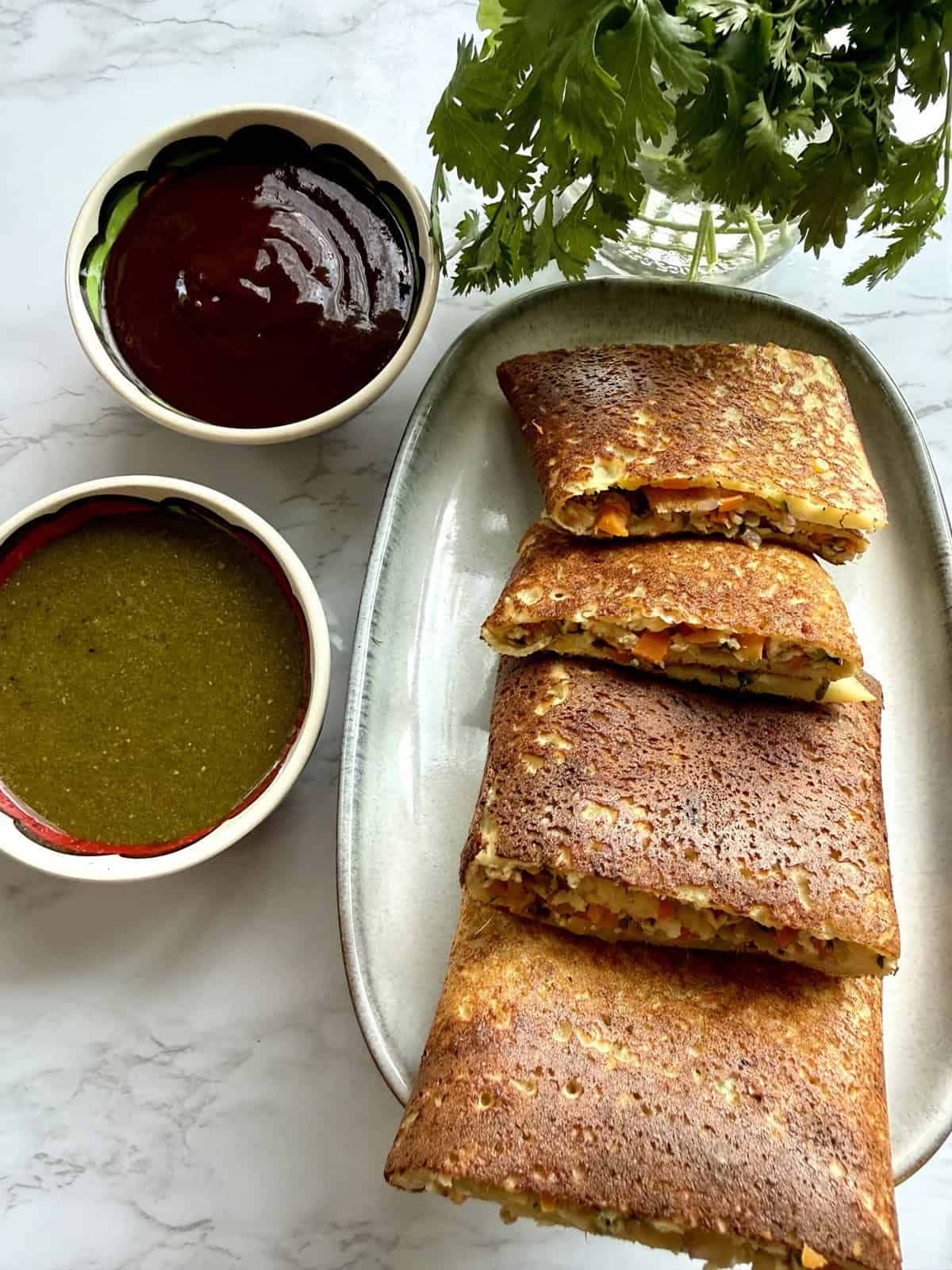 High-protein moong dal cheela with cottage cheese and tofu on a ceramic platter with mint and tamarind chutneys in two bowls on a white marble background.