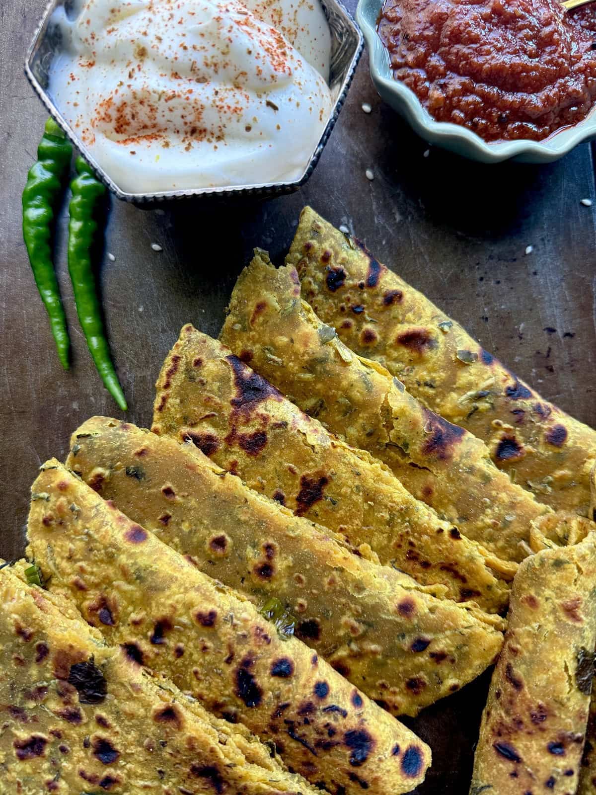 Indian thepla (flatbread) with spicy tomato chutney, yogurt, and green chilies on a dark surface.