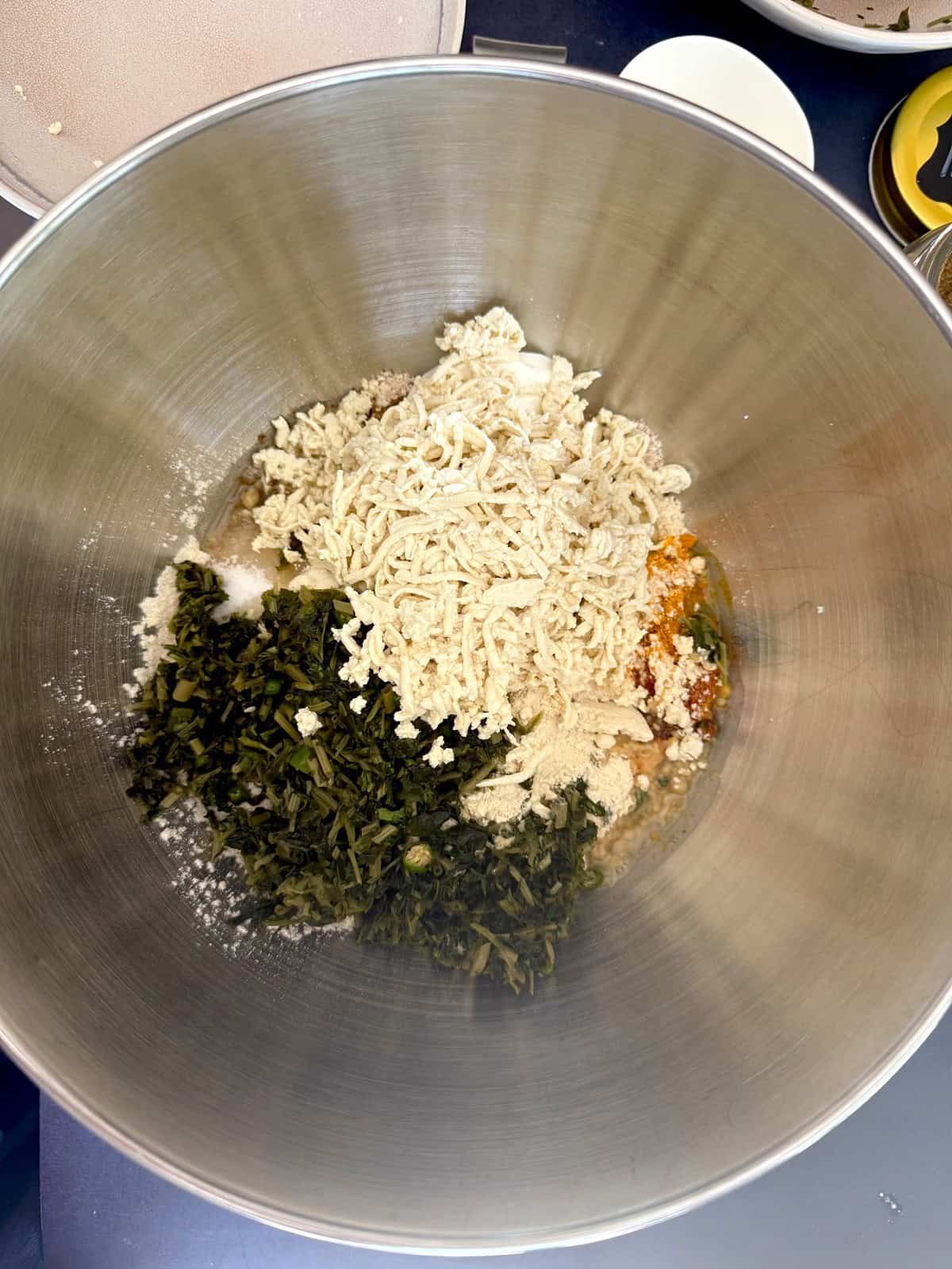 Grated tofu and frozen fenugreek leaves along with other ingredients of high protein tofu theplas in a stainless steel mixing bowl.