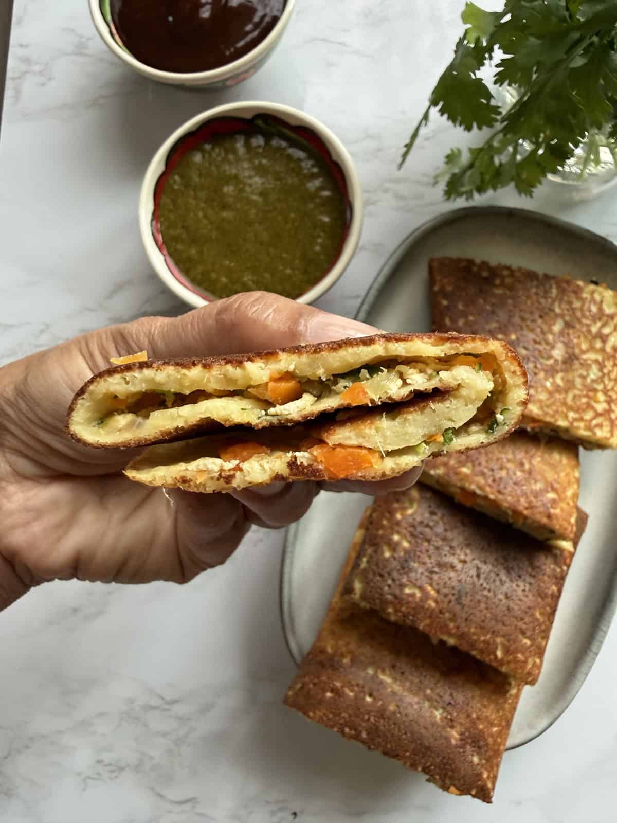 Close up of a slice of High-protein moong dal cheela with cottage cheese and tofu on a ceramic platter with mint and tamarind chutneys in two bowls on a white marble background.