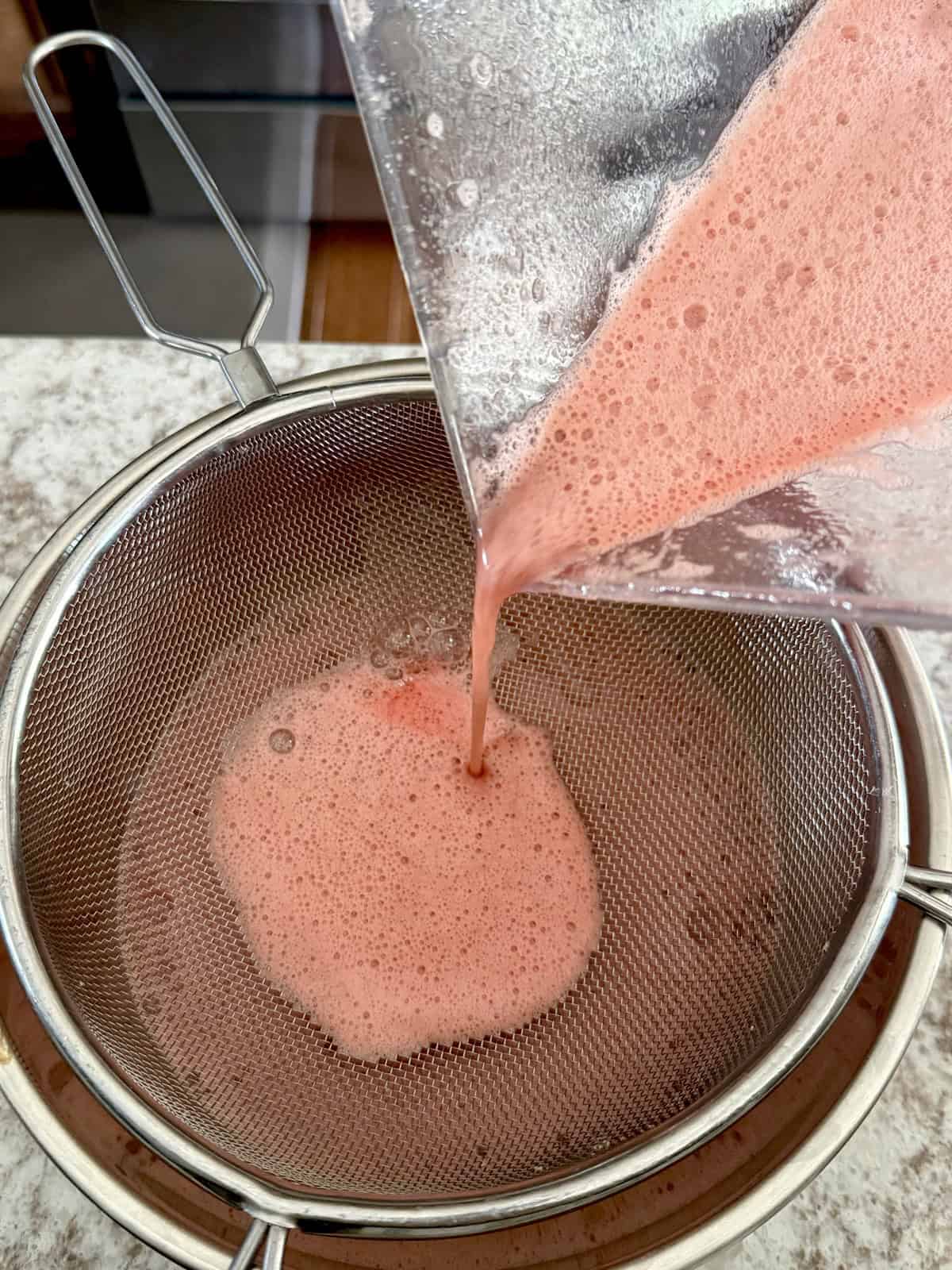 Pouring freshly blended watermelon juice through a fine-mesh metal strainer into a mixing bowl to remove excess pulp for a smooth beverage base.